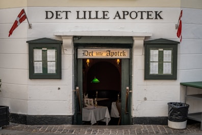 A historic-looking building features an entrance labeled 'Det Lille Apotek' with Danish flags on either side. Two windows display menus on both sides of the entrance, and a cozy interior can be glimpsed with white tablecloths and a green hanging lamp.