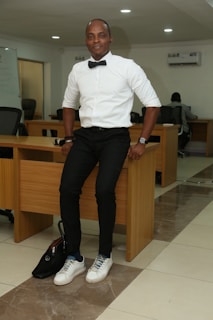 A man in smart casual attire is sitting on the edge of a desk in an office setting. He is wearing a white shirt with a black bow tie, black pants, and white sneakers. The office has wooden desks, black chairs, and a whiteboard in the background. Some people are working at desks.