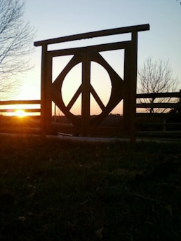 A wooden gate featuring a large peace symbol stands silhouetted against a sunset sky. The sun is low on the horizon, casting an orange glow and long shadows across a grassy field. Leafless trees are visible in the background, framing the scene.