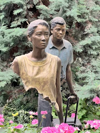 Two life-size outdoor sculptures of a woman and a man surrounded by lush greenery and vibrant pink flowers. The woman wears an abstract, torn top and the man stands slightly behind, pulling a suitcase. Both figures exhibit serious expressions.