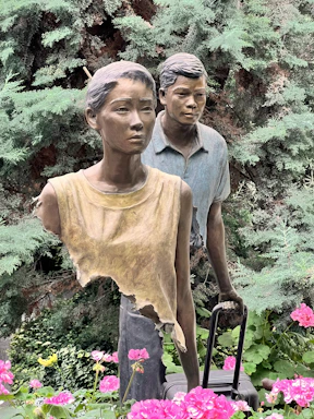 Two life-size outdoor sculptures of a woman and a man surrounded by lush greenery and vibrant pink flowers. The woman wears an abstract, torn top and the man stands slightly behind, pulling a suitcase. Both figures exhibit serious expressions.