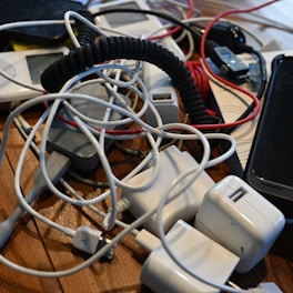 A collection of tangled electronic chargers and cables, including several USB cables, power adapters, and a smartphone, all spread out on a wooden surface.