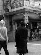 Two people are walking on a street in front of shops with clothing displays. One is wearing a light-colored hoodie and the other a dark coat. Above them, signs are written in multiple languages, including English and Hindi, referencing a welfare association office and a service campaign by OVN Industries Pvt Ltd.