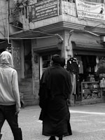 Two people are walking on a street in front of shops with clothing displays. One is wearing a light-colored hoodie and the other a dark coat. Above them, signs are written in multiple languages, including English and Hindi, referencing a welfare association office and a service campaign by OVN Industries Pvt Ltd.