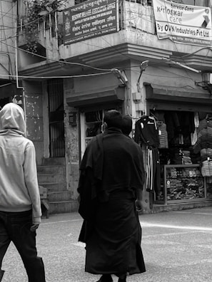 Two people are walking on a street in front of shops with clothing displays. One is wearing a light-colored hoodie and the other a dark coat. Above them, signs are written in multiple languages, including English and Hindi, referencing a welfare association office and a service campaign by OVN Industries Pvt Ltd.