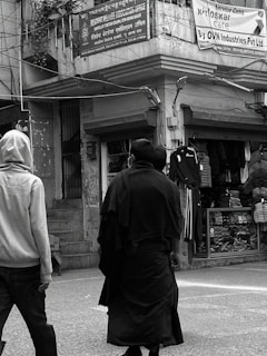 Two people are walking on a street in front of shops with clothing displays. One is wearing a light-colored hoodie and the other a dark coat. Above them, signs are written in multiple languages, including English and Hindi, referencing a welfare association office and a service campaign by OVN Industries Pvt Ltd.