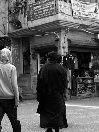 Two people are walking on a street in front of shops with clothing displays. One is wearing a light-colored hoodie and the other a dark coat. Above them, signs are written in multiple languages, including English and Hindi, referencing a welfare association office and a service campaign by OVN Industries Pvt Ltd.