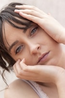 A close-up portrait of a person with light eyes and dark hair, resting their head in their hands. The expression is calm and introspective, with a subtle hint of emotion conveyed through the eyes.