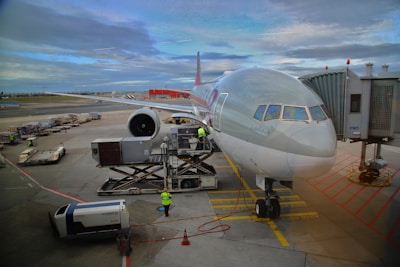 A large commercial airplane is parked at an airport gate with a jet bridge attached to it. Ground crew members in high-visibility vests are working around the aircraft, loading luggage and cargo. The sky is mostly cloudy, and the tarmac is bustling with airport vehicles.