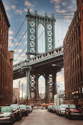 A large suspension bridge spans between two brick buildings, with a view of a city skyline in the background. The street is lined with parked cars, and the architecture includes intricate metalwork on the bridge. The sky is partly cloudy, adding a dramatic effect to the scene.