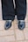 A pair of black leather loafers with gold chain embellishments on each shoe, worn with black pants. The shoes are standing on a light gray cobblestone pavement.