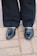 A pair of black leather loafers with gold chain embellishments on each shoe, worn with black pants. The shoes are standing on a light gray cobblestone pavement.