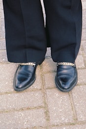 A pair of black leather loafers with gold chain embellishments on each shoe, worn with black pants. The shoes are standing on a light gray cobblestone pavement.