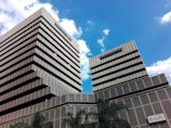 Two modern, multi-story buildings with a geometric design stand tall against a bright blue sky. The buildings have a consistent pattern of horizontal lines and square windows, with tiered levels creating a stepped effect. Palm trees are visible near the base of the buildings, and a sign with the word 'Bogotá' is present on the bottom right corner.
