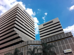 Two modern, multi-story buildings with a geometric design stand tall against a bright blue sky. The buildings have a consistent pattern of horizontal lines and square windows, with tiered levels creating a stepped effect. Palm trees are visible near the base of the buildings, and a sign with the word 'Bogotá' is present on the bottom right corner.