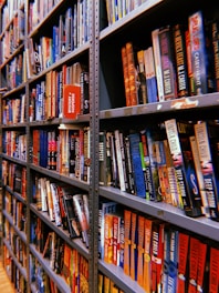 Rows of bookshelves are filled with a variety of colorful books, organized neatly. Some books are titled visibly with genres like mystery and suspense, and they are packed tightly together on metal shelves.