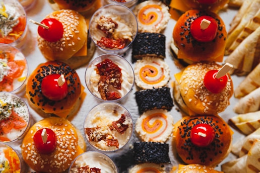 An assortment of appetizing finger foods is displayed, including mini burgers with sesame and black seeds topped with cherry tomatoes, cups of creamy dips garnished with dried tomatoes, sushi rolls with orange and black fillings, and various rolled crepes or wraps. The arrangement is colorful and appears to be a buffet or party platter.