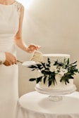 A close-up of hands cutting a wedding cake adorned with fresh flowers and greenery.