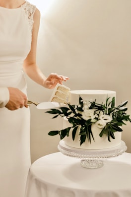 A close-up of hands cutting a wedding cake adorned with fresh flowers and greenery.
