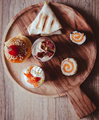 A wooden round serving board displays an assortment of small appetizers. There is a mini burger topped with sesame seeds and a cherry, a wrapped sandwich, a piece of rolled sushi, and two small glass cups filled with creamy desserts garnished with fruit and toppings.