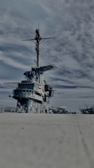 A large naval warship on a concrete surface under a partly cloudy sky. The ship displays multiple decks and an extensive array of communication equipment on its mast. Several other aircraft are visible in the distance on the right side.