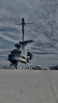 A large naval warship on a concrete surface under a partly cloudy sky. The ship displays multiple decks and an extensive array of communication equipment on its mast. Several other aircraft are visible in the distance on the right side.