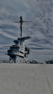 A large naval warship on a concrete surface under a partly cloudy sky. The ship displays multiple decks and an extensive array of communication equipment on its mast. Several other aircraft are visible in the distance on the right side.