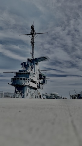 A large naval warship on a concrete surface under a partly cloudy sky. The ship displays multiple decks and an extensive array of communication equipment on its mast. Several other aircraft are visible in the distance on the right side.
