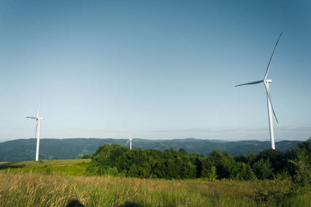 Three large wind turbines stand tall on a grassy field, set against a backdrop of distant mountains and a clear blue sky. The landscape features lush greenery with rolling hills in the distance, and the turbines are evenly spaced, each one sleek and modern in design.