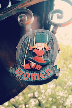 A rustic wooden sign adorned with a whimsical illustration of a blonde-haired character in a red outfit and hat. The sign features decorative scrollwork at the top and the word 'women' in bold, colorful letters.