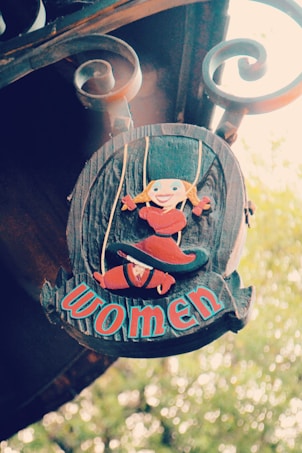 A rustic wooden sign adorned with a whimsical illustration of a blonde-haired character in a red outfit and hat. The sign features decorative scrollwork at the top and the word 'women' in bold, colorful letters.