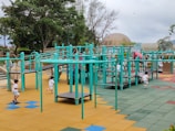 A vibrant playground setting features multiple children engaging in active play. The playground structure is comprised of turquoise bars and platforms, with slides and climbing elements. The surface is covered in colorful interlocking tiles, primarily in shades of yellow, blue, green, and red. Surrounding the play area are lush green trees and a dome-shaped structure in the background.