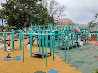 A vibrant playground setting features multiple children engaging in active play. The playground structure is comprised of turquoise bars and platforms, with slides and climbing elements. The surface is covered in colorful interlocking tiles, primarily in shades of yellow, blue, green, and red. Surrounding the play area are lush green trees and a dome-shaped structure in the background.