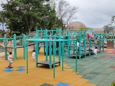 A vibrant playground setting features multiple children engaging in active play. The playground structure is comprised of turquoise bars and platforms, with slides and climbing elements. The surface is covered in colorful interlocking tiles, primarily in shades of yellow, blue, green, and red. Surrounding the play area are lush green trees and a dome-shaped structure in the background.