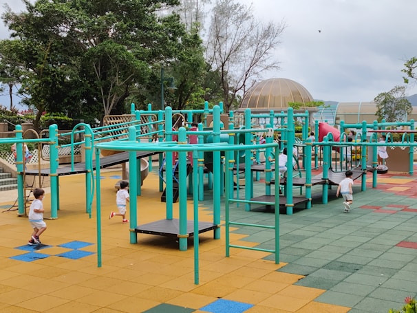 A vibrant playground setting features multiple children engaging in active play. The playground structure is comprised of turquoise bars and platforms, with slides and climbing elements. The surface is covered in colorful interlocking tiles, primarily in shades of yellow, blue, green, and red. Surrounding the play area are lush green trees and a dome-shaped structure in the background.
