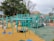 A vibrant playground setting features multiple children engaging in active play. The playground structure is comprised of turquoise bars and platforms, with slides and climbing elements. The surface is covered in colorful interlocking tiles, primarily in shades of yellow, blue, green, and red. Surrounding the play area are lush green trees and a dome-shaped structure in the background.