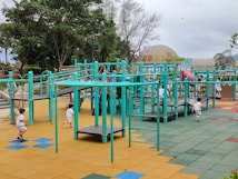 A vibrant playground setting features multiple children engaging in active play. The playground structure is comprised of turquoise bars and platforms, with slides and climbing elements. The surface is covered in colorful interlocking tiles, primarily in shades of yellow, blue, green, and red. Surrounding the play area are lush green trees and a dome-shaped structure in the background.