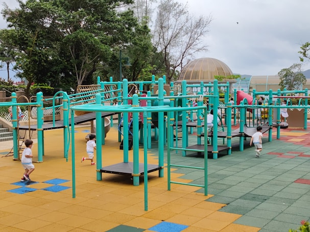 A vibrant playground setting features multiple children engaging in active play. The playground structure is comprised of turquoise bars and platforms, with slides and climbing elements. The surface is covered in colorful interlocking tiles, primarily in shades of yellow, blue, green, and red. Surrounding the play area are lush green trees and a dome-shaped structure in the background.
