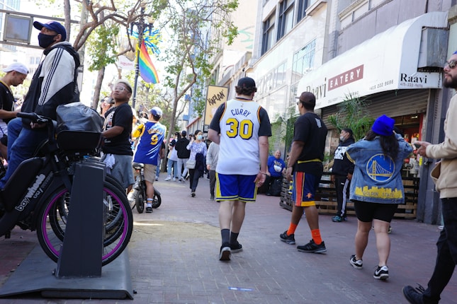A bustling urban street with several people walking and standing. Many are wearing sports jerseys, prominently featuring blue and yellow colors. The scene includes a bike with a vivid purple wheel parked nearby, and a rainbow flag hangs in the background. The street is lined with buildings, including a visible eatery.
