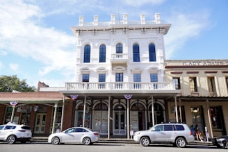 A historic three-story brick building with arched windows and a decorative balcony stands prominently. It is flanked by smaller, attached structures with wooden facades. Three parked cars line the street outside the buildings. Patriotic buntings are draped along the balcony railing, adding a festive touch. The sky is clear with a few scattered clouds.