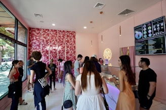 A lively boba tea shop interior with customers lining up at the counter. The decor includes a floral wall with pink hues and neon signage, creating a trendy and vibrant atmosphere. Large menu boards display various drink options. Multiple people are engaged in conversation or waiting to order.