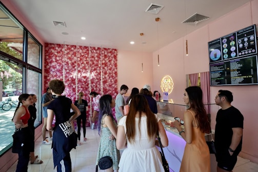 A lively boba tea shop interior with customers lining up at the counter. The decor includes a floral wall with pink hues and neon signage, creating a trendy and vibrant atmosphere. Large menu boards display various drink options. Multiple people are engaged in conversation or waiting to order.