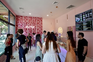 A lively boba tea shop interior with customers lining up at the counter. The decor includes a floral wall with pink hues and neon signage, creating a trendy and vibrant atmosphere. Large menu boards display various drink options. Multiple people are engaged in conversation or waiting to order.