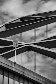 An architectural structure featuring an arched steel bridge with intersecting beams against a cloudy sky. A speed limit sign indicating 35 is visible in the foreground, attached to a concrete segment of the bridge.