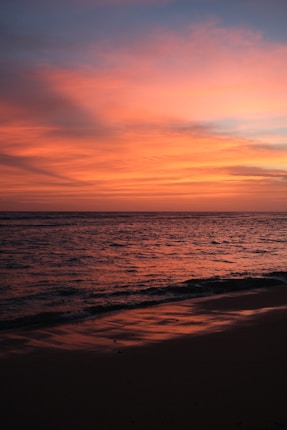 A serene beach with gentle waves lapping the shore, under a dramatic sunset sky filled with shades of orange, pink, and violet clouds.