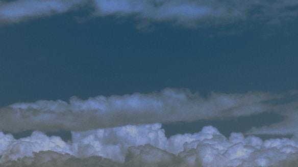 Fluffy white clouds gather in the lower part of the image against a backdrop of deep blue sky. The clouds appear thick and voluminous, capturing the play of light and shadow across their surfaces.
