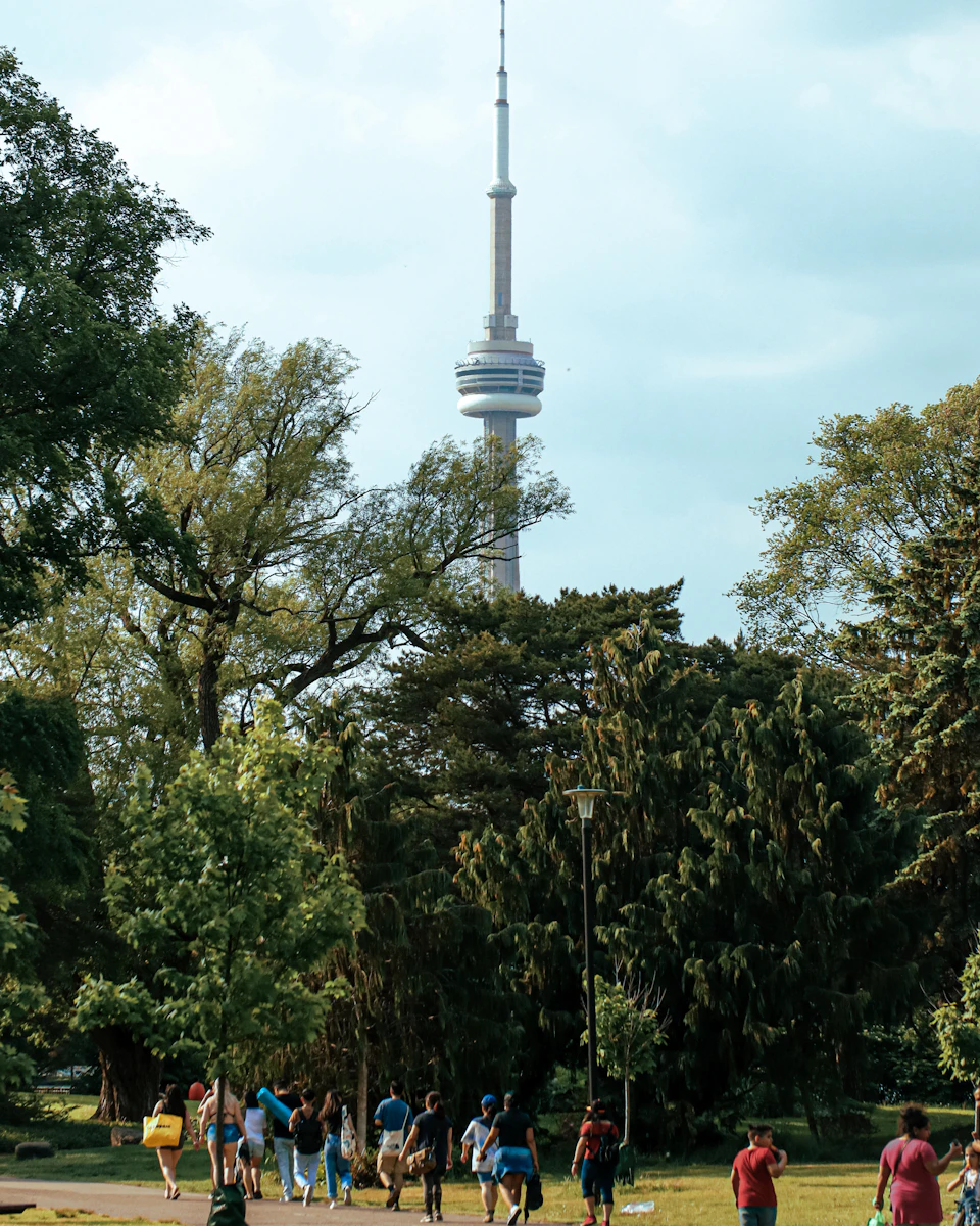 The image depicts a group of people walking through a park, with some individuals carrying backpacks and handbags. The park is lush with trees, providing a natural backdrop to the scene. In the distance, a tall white tower can be seen against the sky.