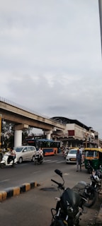 A busy urban street scene featuring a variety of vehicles, including cars, auto rickshaws, motorbikes, and a bus. A prominent elevated metro track runs above the road, adjacent to a metro station. The sky is overcast, suggesting possible cloud cover.