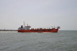 A large red cargo ship is sailing on a calm sea. The ship has various equipment and structures on its deck, including a crane. The horizon is visible in the background under a clear, pale sky.