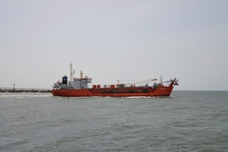 A large red cargo ship is sailing on a calm sea. The ship has various equipment and structures on its deck, including a crane. The horizon is visible in the background under a clear, pale sky.
